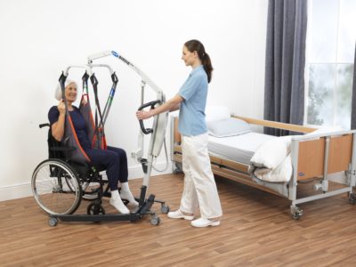 Eine Pflegekraft hilft einer älteren Dame mithilfe eines Patientenlifters beim Transfer aus ihrem Rollstuhl. Sie haben Blickkontakt. 