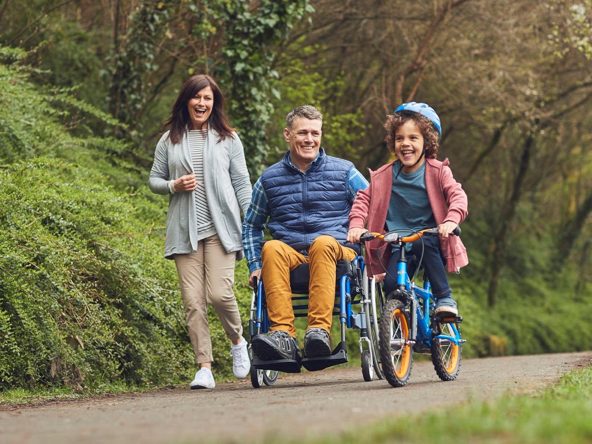 Ein junger, fröhlicher Mann fährt mit seinem Aktivrollstuhl MyOn HC Rigid durch den Park. Mit dabei sind seine Frau und sein Sohn auf dem Fahrrad.
