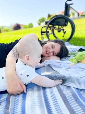 Eine Frau liegt mit ihrem Baby auf einer Decke, auf einer Wiese. Im Hintergrund steht ihr Rollstuhl.